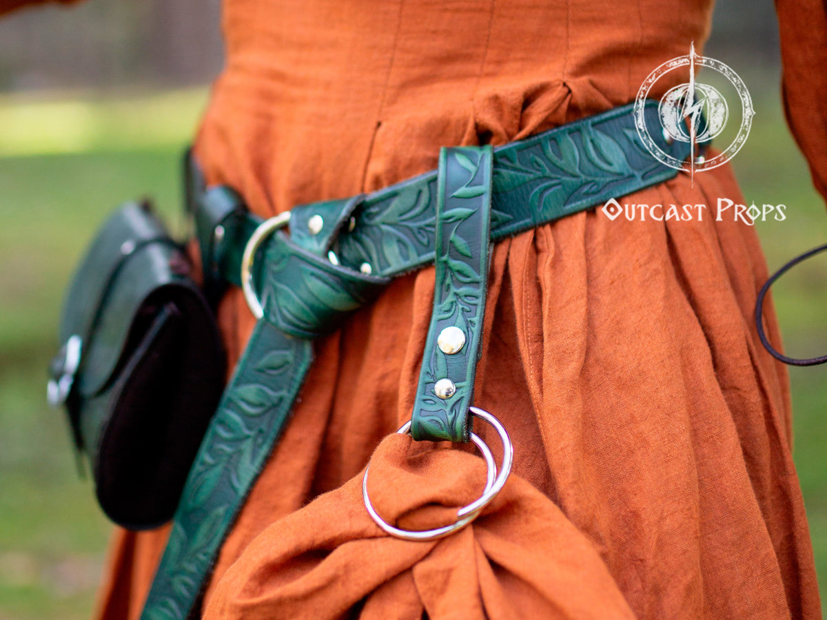 A handcrafted green leather skirt hike with a leaf motif, tooling, and a double O-ring system attached to a brown belt, worn over a character's costume. Medieval Floral Leather Skirt Hike Ring LARP Ren Faire Cosplay Accessory for Fantasy Outfits Skirt Hiker Renaissance Fair Costume Accessory