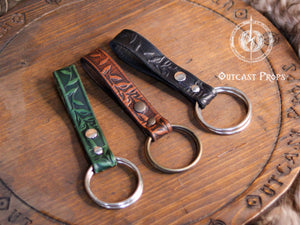 Three Skirt Hikes with metal rings. The skirt hikes  are brown, green, and black with a floral design. The skirt hikes are displayed on a wooden surface with the text 'Outcast Props'. Medieval Floral Leather Skirt Hike Ring LARP Ren Faire Cosplay Accessory for Fantasy Outfits Skirt Hiker Renaissance Fair Costume Accessory