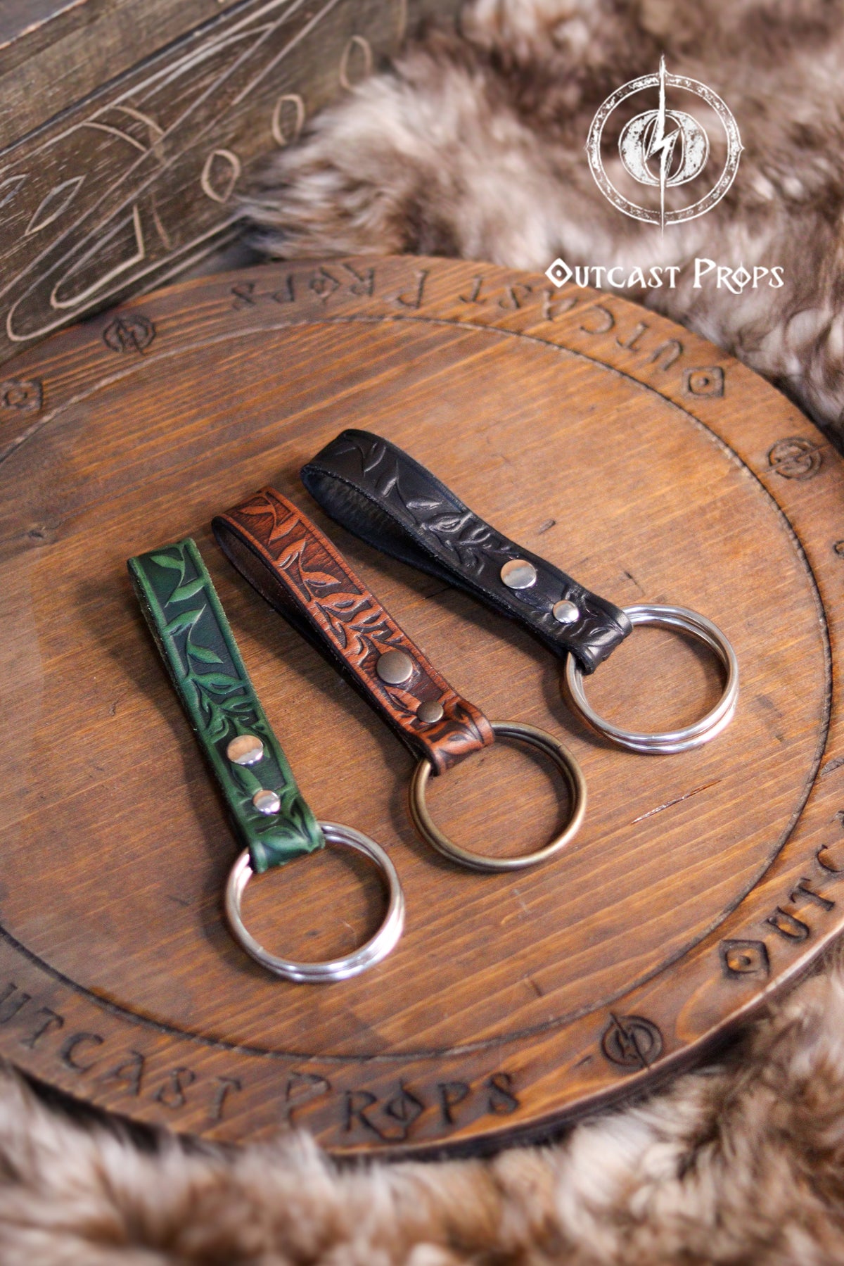 Three Skirt Hike with metal rings. The skirt hikes  are brown, green, and black with a floral design. The skirt hikes are displayed on a wooden surface with the text 'Outcast Props'. Medieval Floral Leather Skirt Hike Ring LARP Ren Faire Cosplay Accessory for Fantasy Outfits Skirt Hiker Renaissance Fair Costume Accessory