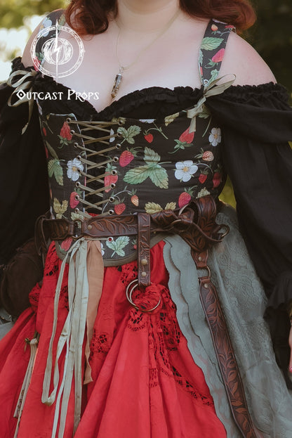 A close-up of a person wearing a medieval-style costume dress with a floral corset bodice, black off-shoulder sleeves, a brown leather-look belt, and a red skirt with gray fabric accents. The background is blurred green foliage.