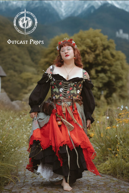 A woman stands barefoot on a stone path in a grassy outdoor setting with mountains in the background, wearing an ornate medieval-style costume dress. The dress has a black bodice with floral embroidery, a red and black layered skirt, and dark sleeves.
