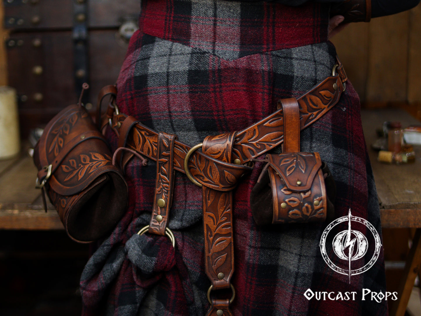 A close view of a brown floral leather skirt hike attached to a wide belt worn over a red and gray plaid wool skirt. The handcrafted accessory features leaf embossing, a double O-ring system and snap strap, lifting a section of the fabric to create a layered fantasy look. Matching pouches and holsters hang from the belt, forming a cohesive Renaissance faire or LARP outfit suited for elves, rangers, witches or medieval characters who want both style and practical carry options.