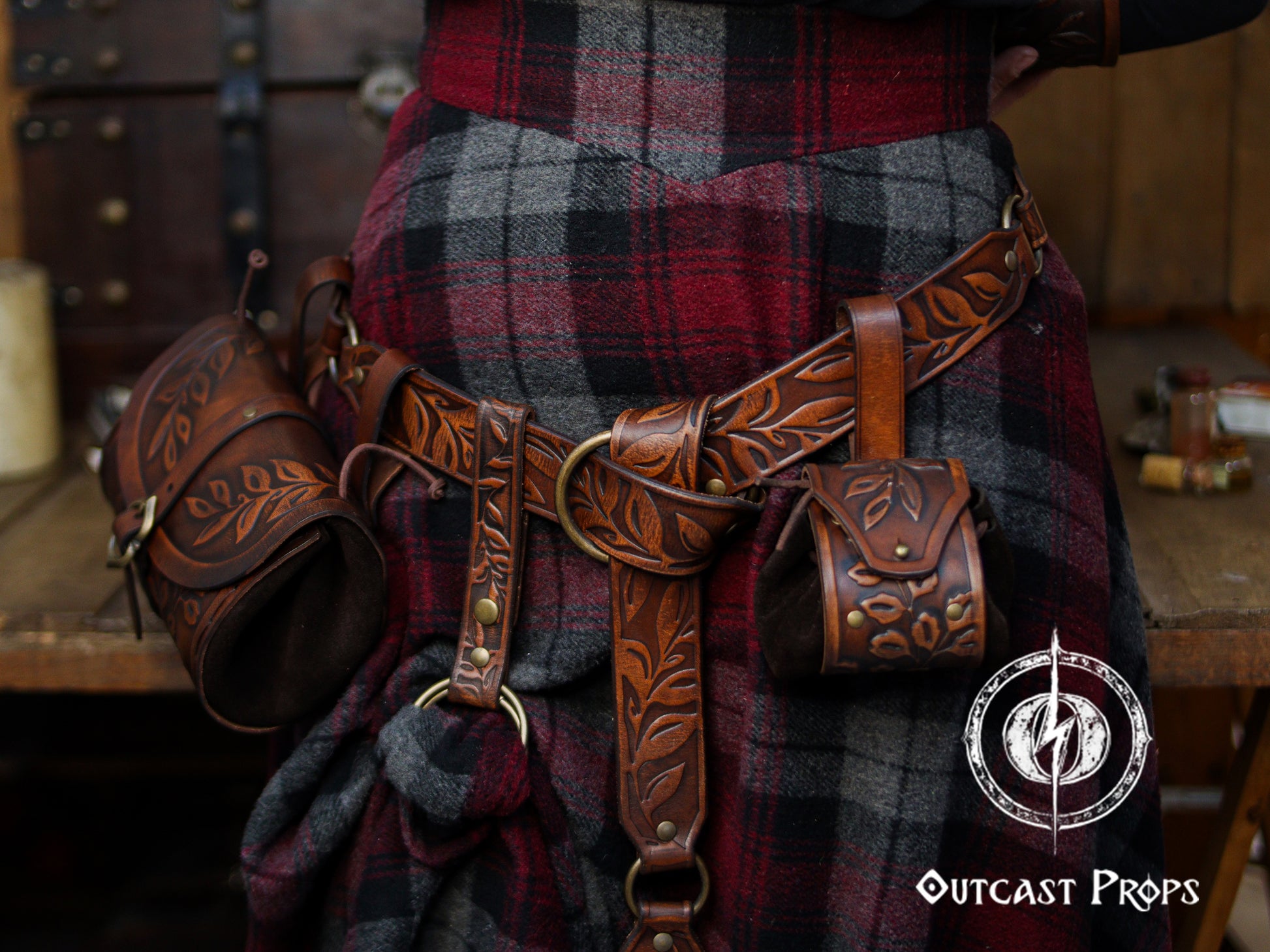 A close view of a brown floral leather skirt hike attached to a wide belt worn over a red and gray plaid wool skirt. The handcrafted accessory features leaf embossing, a double O-ring system and snap strap, lifting a section of the fabric to create a layered fantasy look. Matching pouches and holsters hang from the belt, forming a cohesive Renaissance faire or LARP outfit suited for elves, rangers, witches or medieval characters who want both style and practical carry options.