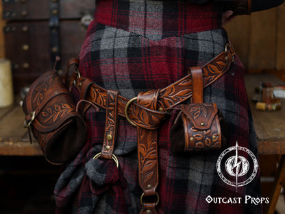 A close view of a brown floral leather skirt hike attached to a wide belt worn over a red and gray plaid wool skirt. The handcrafted accessory features leaf embossing, a double O-ring system and snap strap, lifting a section of the fabric to create a layered fantasy look. Matching pouches and holsters hang from the belt, forming a cohesive Renaissance faire or LARP outfit suited for elves, rangers, witches or medieval characters who want both style and practical carry options.