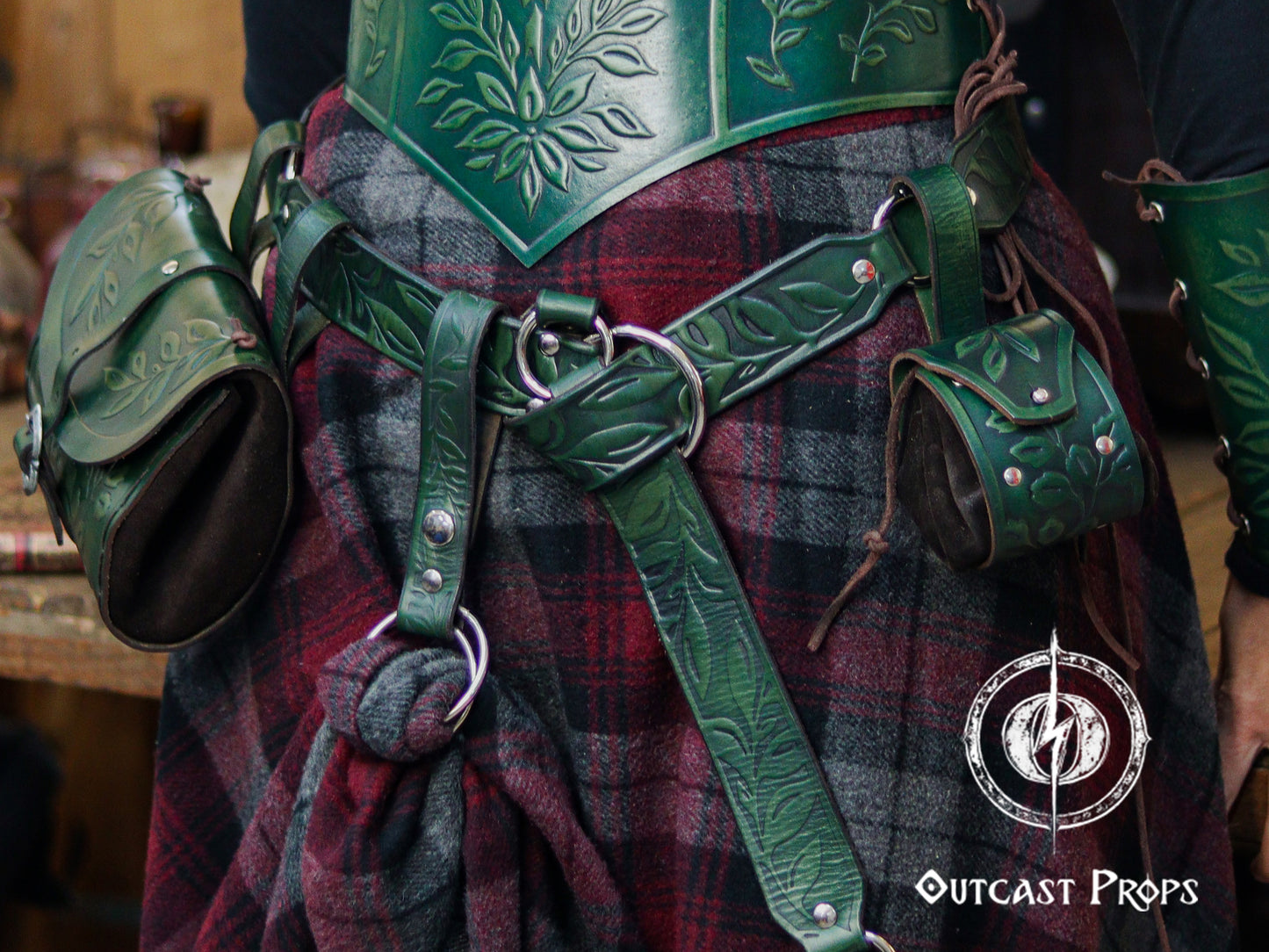 A green version of the floral leather skirt hike is shown clipped to a belt over a thick plaid wool skirt, paired with matching green tooled leather armor and pouches. The embossed leaf pattern, double O-ring system and snap closure allow the wearer to gather fabric and secure it for movement or style. Ideal for LARP, Renaissance faire and fantasy costumes, the accessory adds charm while supporting pouches, tankards or potion holders as part of an immersive character outfit.