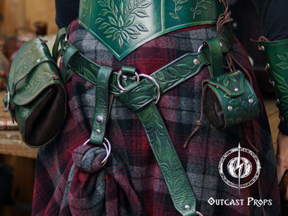 A green version of the floral leather skirt hike is shown clipped to a belt over a thick plaid wool skirt, paired with matching green tooled leather armor and pouches. The embossed leaf pattern, double O-ring system and snap closure allow the wearer to gather fabric and secure it for movement or style. Ideal for LARP, Renaissance faire and fantasy costumes, the accessory adds charm while supporting pouches, tankards or potion holders as part of an immersive character outfit.