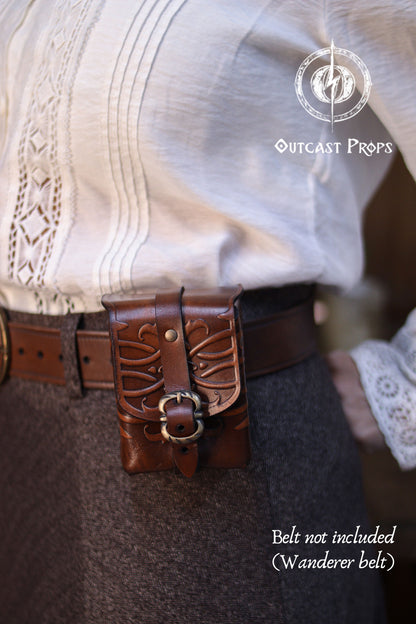 Close-up of an ornated medieval leather tobacco bag attached to a belt, showing embossed floral patterns, hand-dyed finish, and solid brass buckle. Crafted from premium vegetable-tanned leather, this compact belt pouch is ideal for everyday use, tabletop card games, or as a thoughtful handmade leather gift for fantasy, steampunk, or Renaissance fair enthusiasts. Close-up of a belt with decorative buckle worn by a person, with 'Outcast Props' branding.