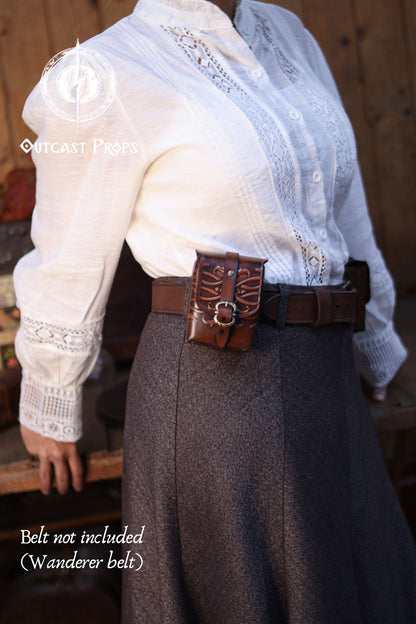 Side view of an ornated leather belt pouch worn at the waist, highlighting its clean silhouette and secure closure. Handmade from vegetable-tanned leather using traditional techniques, this medieval tobacco bag works as a card deck holder, fantasy costume accessory, or refined everyday pouch. A versatile leather gift combining practicality and artisan craftsmanship. Close-up of a person wearing a white blouse with lace details and a brown belt, standing against a wooden background.