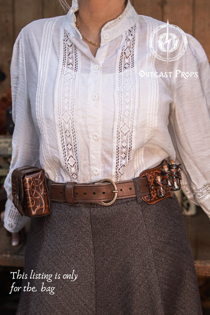 Front view of an ornated leather tobacco bag worn on a belt, crafted from vegetable-tanned leather with hand-tooled embossing and aged finish. Designed for carrying cards, tobacco, or small essentials, this medieval leather belt pouch is perfect as a handmade gift, fantasy accessory, or elegant everyday leather bag for those who value craftsmanship and style. Person wearing a white embroidered shirt with a brown leather belt featuring metal buckles.