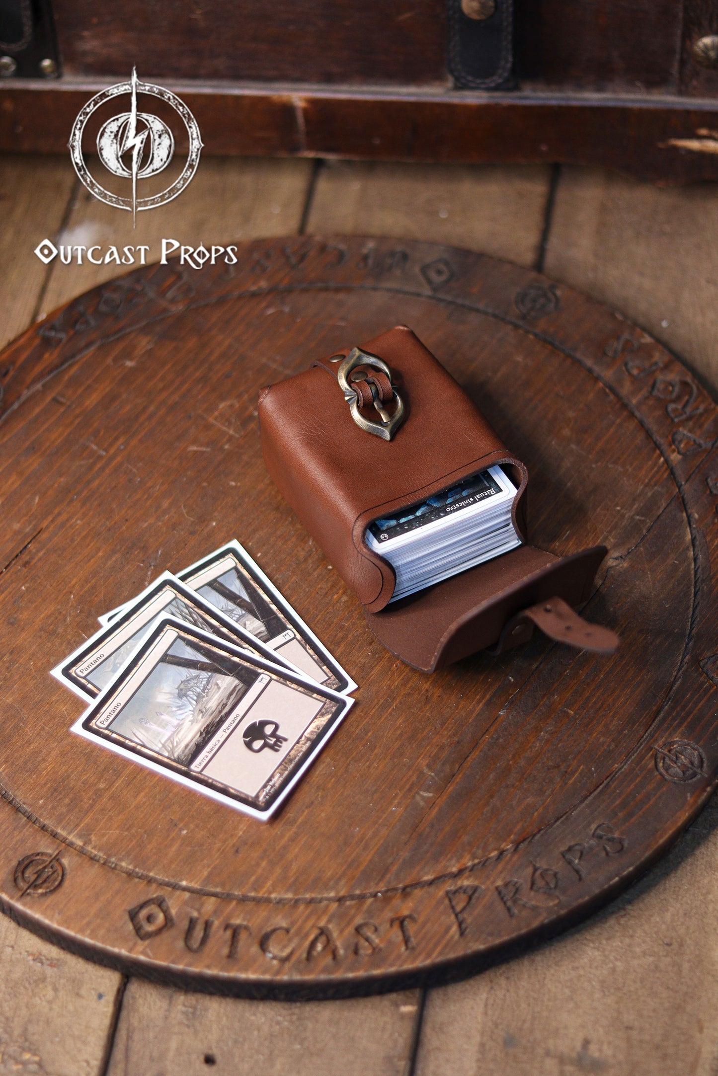 Handcrafted medieval leather belt pouch shown open on a wooden surface, made from premium vegetable tanned leather with a warm brown finish and brass buckle closure. The pouch is displayed as a compact deck bag holding trading cards, suitable for Magic The Gathering, Pokémon TCG, and Arkham Horror card games. Its sturdy structure protects cards while maintaining a historical tavern inspired look. Designed to attach to belts up to 5 cm wide, this leather belt pouch also works as a coin bag, tobacco pouch, di