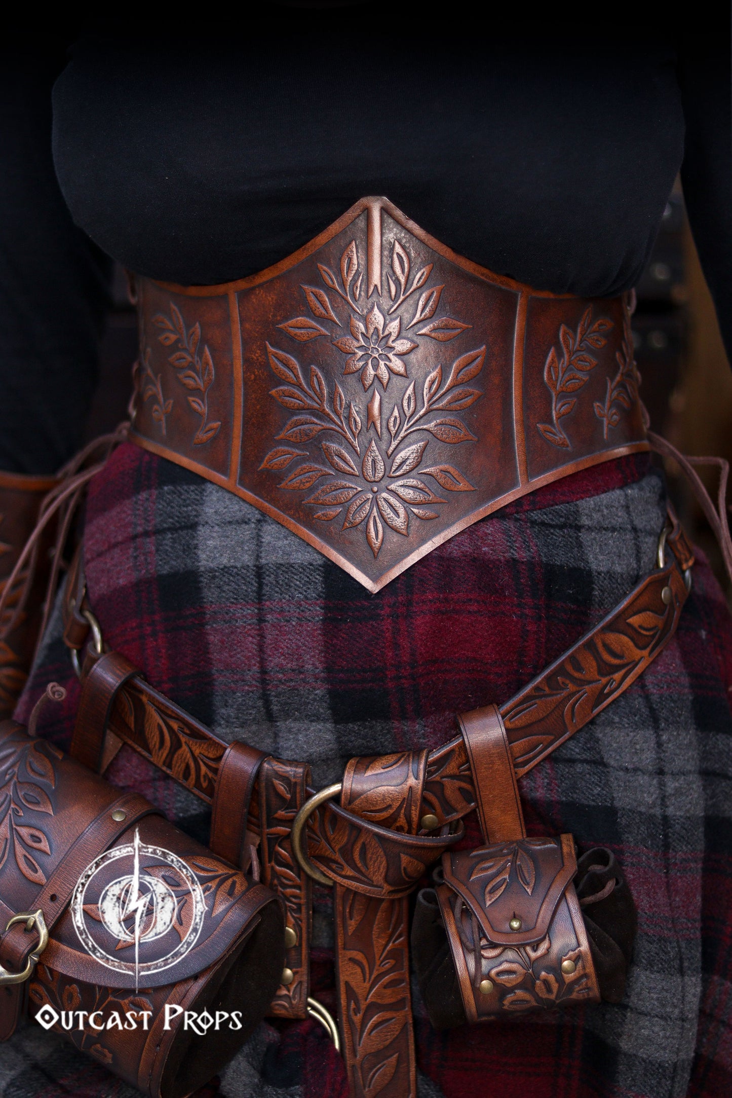 Floral embossed leather underbust corset in aged brown, crafted from premium vegetable tanned leather and worn as part of a Renaissance Faire outfit. The image highlights the detailed leaf carvings, ideal for elves, witches, herbalists or nobles at LARP or renfaire events. Below the corset, a matching belt and coin pouches complete the elegant medieval fantasy look while keeping essentials close during immersive roleplay.