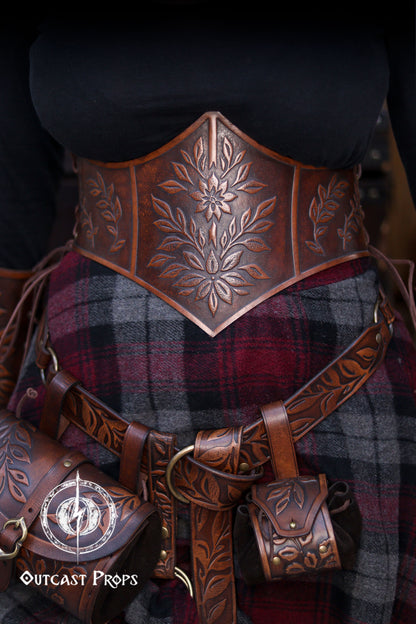 Floral embossed leather underbust corset in aged brown, crafted from premium vegetable tanned leather and worn as part of a Renaissance Faire outfit. The image highlights the detailed leaf carvings, ideal for elves, witches, herbalists or nobles at LARP or renfaire events. Below the corset, a matching belt and coin pouches complete the elegant medieval fantasy look while keeping essentials close during immersive roleplay.