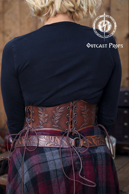 Back view of a floral leather underbust corset belt in aged brown worn over a plaid fantasy skirt during a renaissance faire style setting. The lacing runs across the entire back, showing how the corset adjusts naturally to fit S to L bodies. Embossed leaf patterns highlight the handcrafted look. Ideal for elven cosplay, herbalist outfits, witchcore clothing, LARP costumes and medieval inspired fashion. Outcast Props logo visible in the corner.