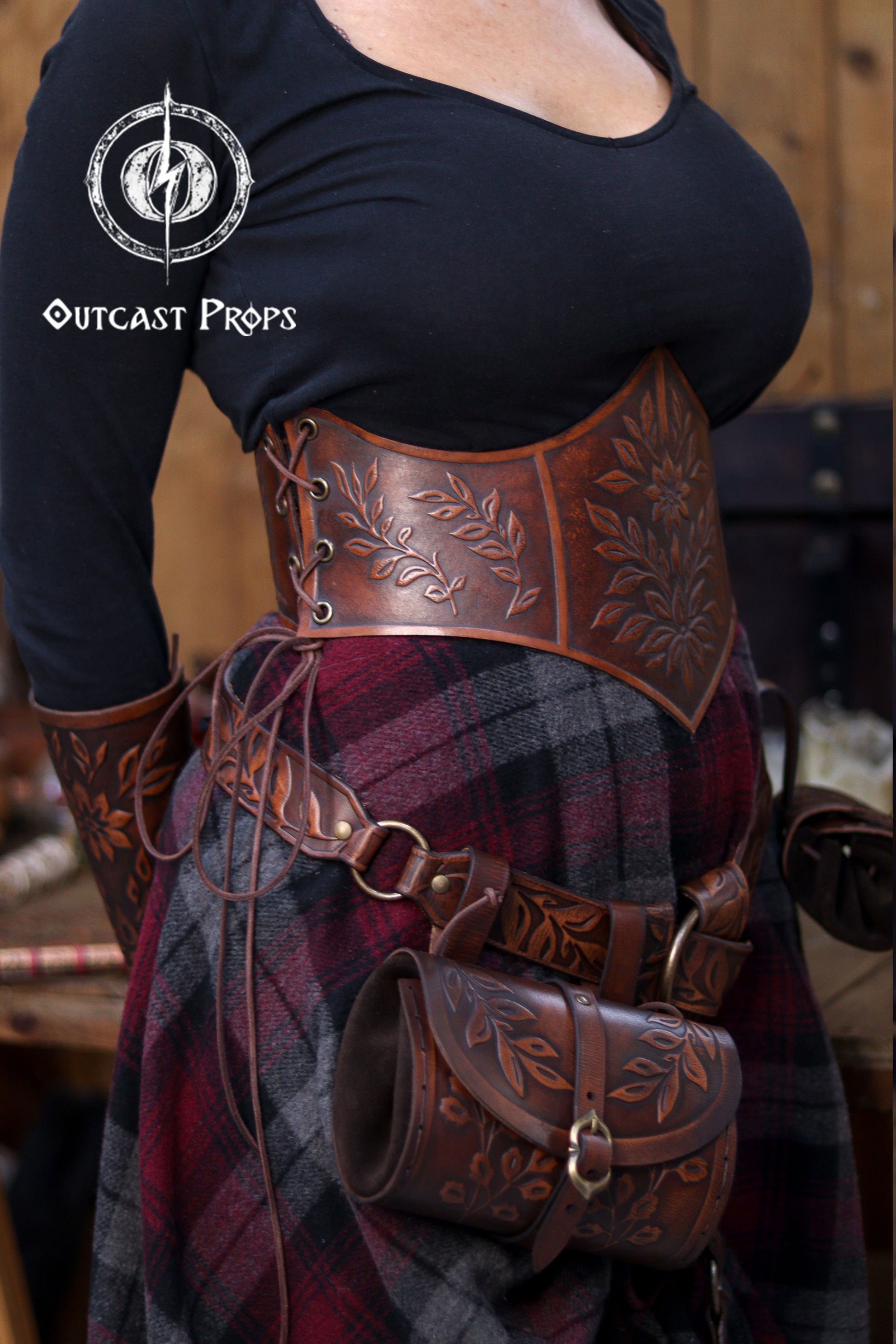 Side view of a model wearing an aged brown floral leather underbust corset belt paired with matching accessories from the same floral set. The embossed leaves and artisan aging give it a rich renaissance faire aesthetic. The corset cinches the waist beautifully, perfect for elven characters, witches, herbalists, fantasy cosplay and LARP costumes. The belt pouches and skirt hike add practical medieval flair.