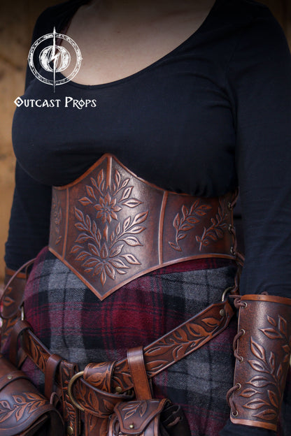 Close side view of the floral leather underbust corset belt highlighting the botanical embossing and warm aged brown tones. The lacing on the side shows the adjustable nature of the corset, making it suitable for LARP costumes, elven outfits, witchcore fashion and renaissance faire clothing. Matching accessories and fantasy skirt complete the handcrafted look. Outcast Props logo appears in the corner.