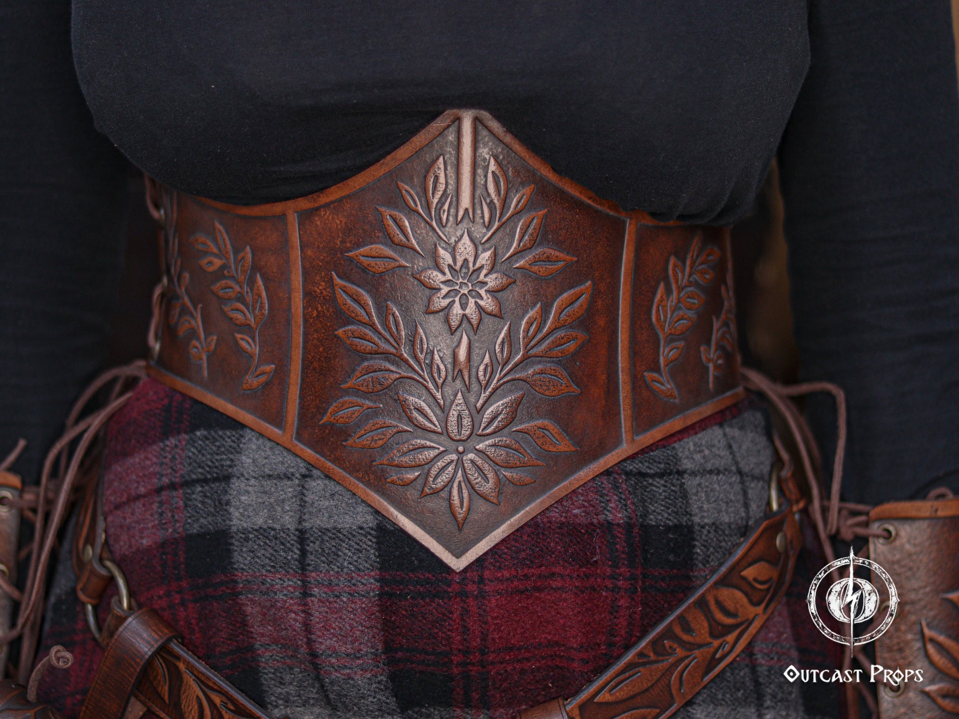 Front view of the floral leather underbust corset belt showing the detailed embossing and elegant curved shape that defines the waist and enhances the bust. The aged brown leather pairs with a plaid fantasy skirt and matching floral belt accessories. Ideal for renaissance faire outfits, elven cosplay, witchcore style, herbalist characters and LARP costumes. The handcrafted finish gives it a cinematic fantasy look.