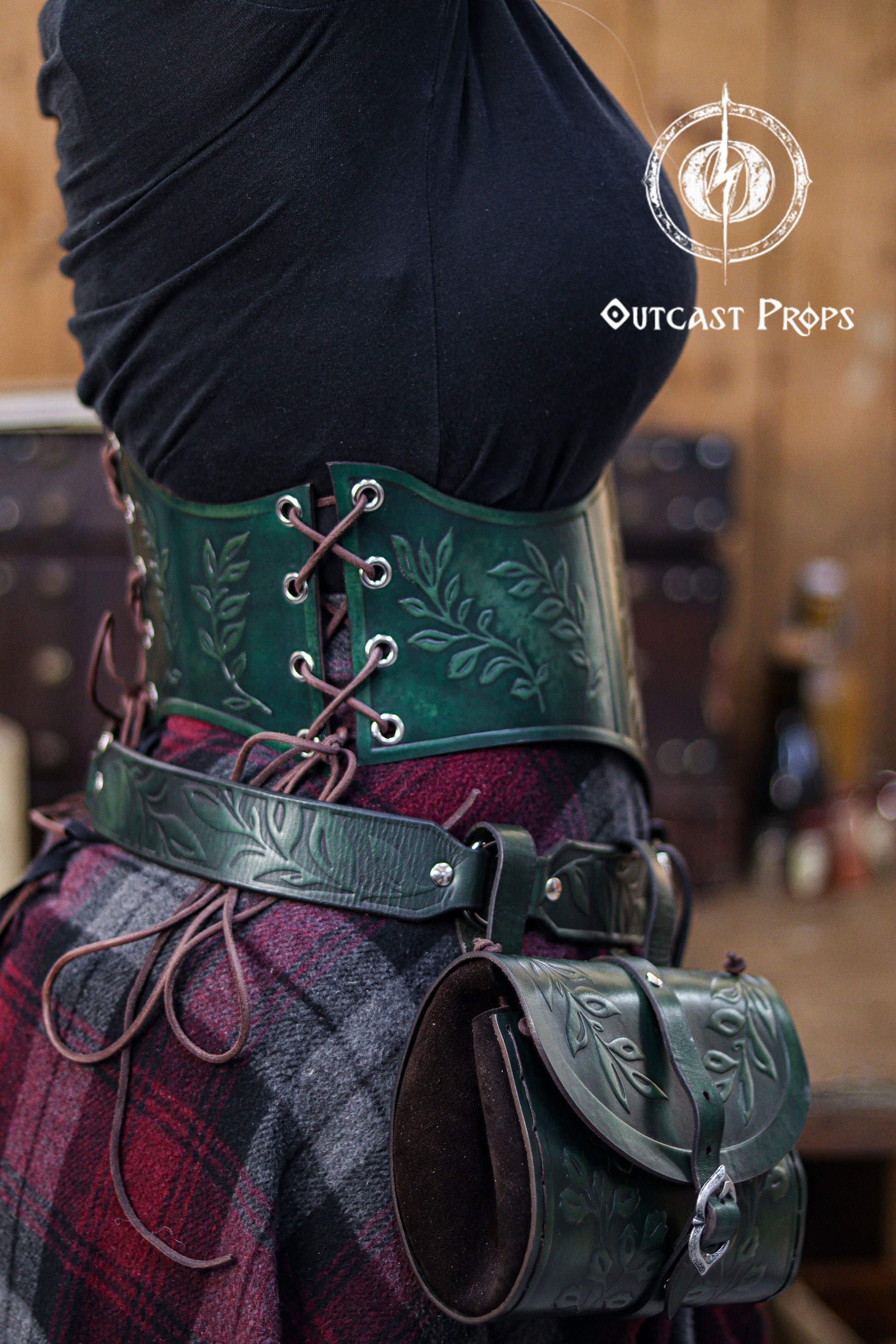 Side view of a model wearing the green floral leather underbust corset belt from Outcast Props, paired with a matching embossed belt pouch. The deep emerald color and botanical embossing create a strong elven and herbalist aesthetic ideal for renaissance faire looks, witchcore outfits, fantasy cosplay and LARP costumes. The lacing on the sides shows the adjustable fit, and the aged finish highlights the handcrafted, cinematic style.