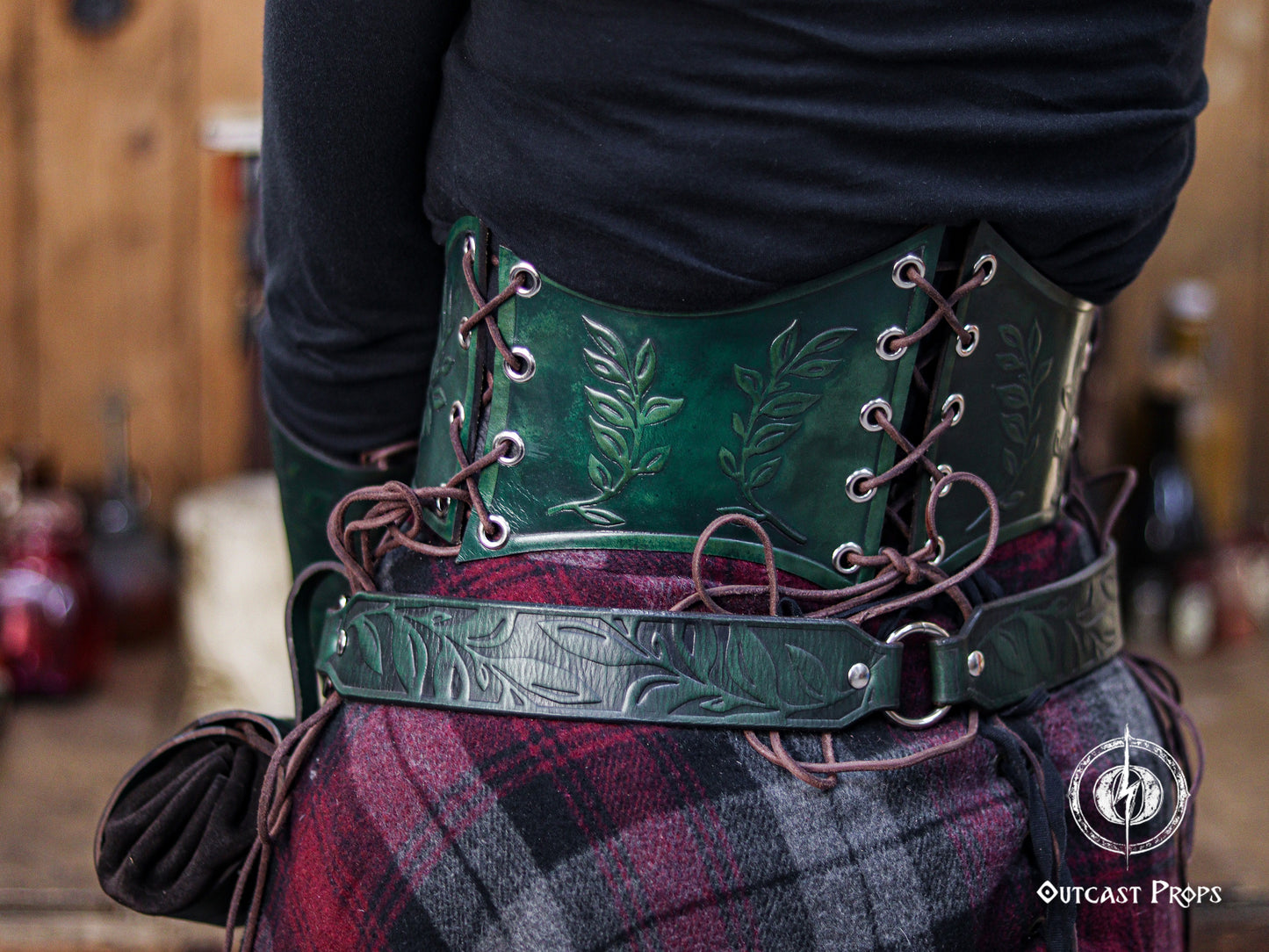 Back angled view of the green floral underbust corset belt, featuring botanical embossing and rich emerald tones. The lacing system enhances the adjustable fit, making it suitable for elven cosplay, renaissance faire characters, witchcore fashion and herbalist inspired outfits. The matching embossed belt adds to the coordinated fantasy look. The image captures the handcrafted aging techniques for a natural, magical, forest themed style.
