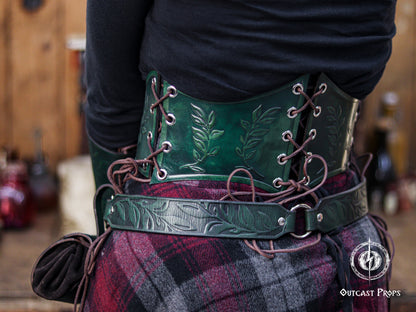 Back angled view of the green floral underbust corset belt, featuring botanical embossing and rich emerald tones. The lacing system enhances the adjustable fit, making it suitable for elven cosplay, renaissance faire characters, witchcore fashion and herbalist inspired outfits. The matching embossed belt adds to the coordinated fantasy look. The image captures the handcrafted aging techniques for a natural, magical, forest themed style.