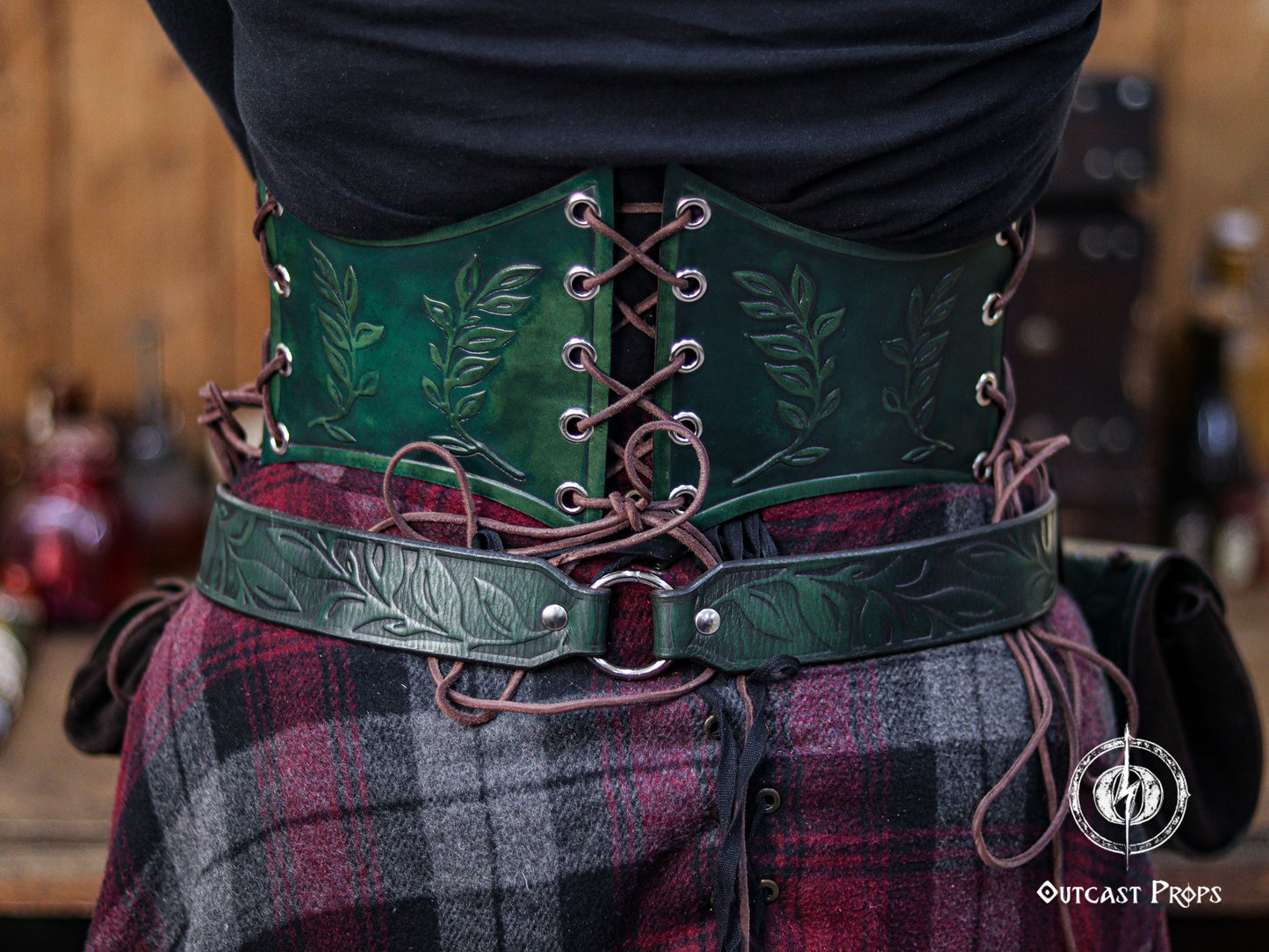 Back view of the green floral leather underbust corset belt showing the full lacing pattern and detailed leaf embossing. The dark emerald dye and handcrafted finish evoke an elven, druidic and herbalist theme ideal for LARP costumes, fantasy events, renaissance faires and witchcore fashion. The matching belt creates a cohesive outfit with cinematic textures typical of handcrafted leather accessories by Outcast Props.