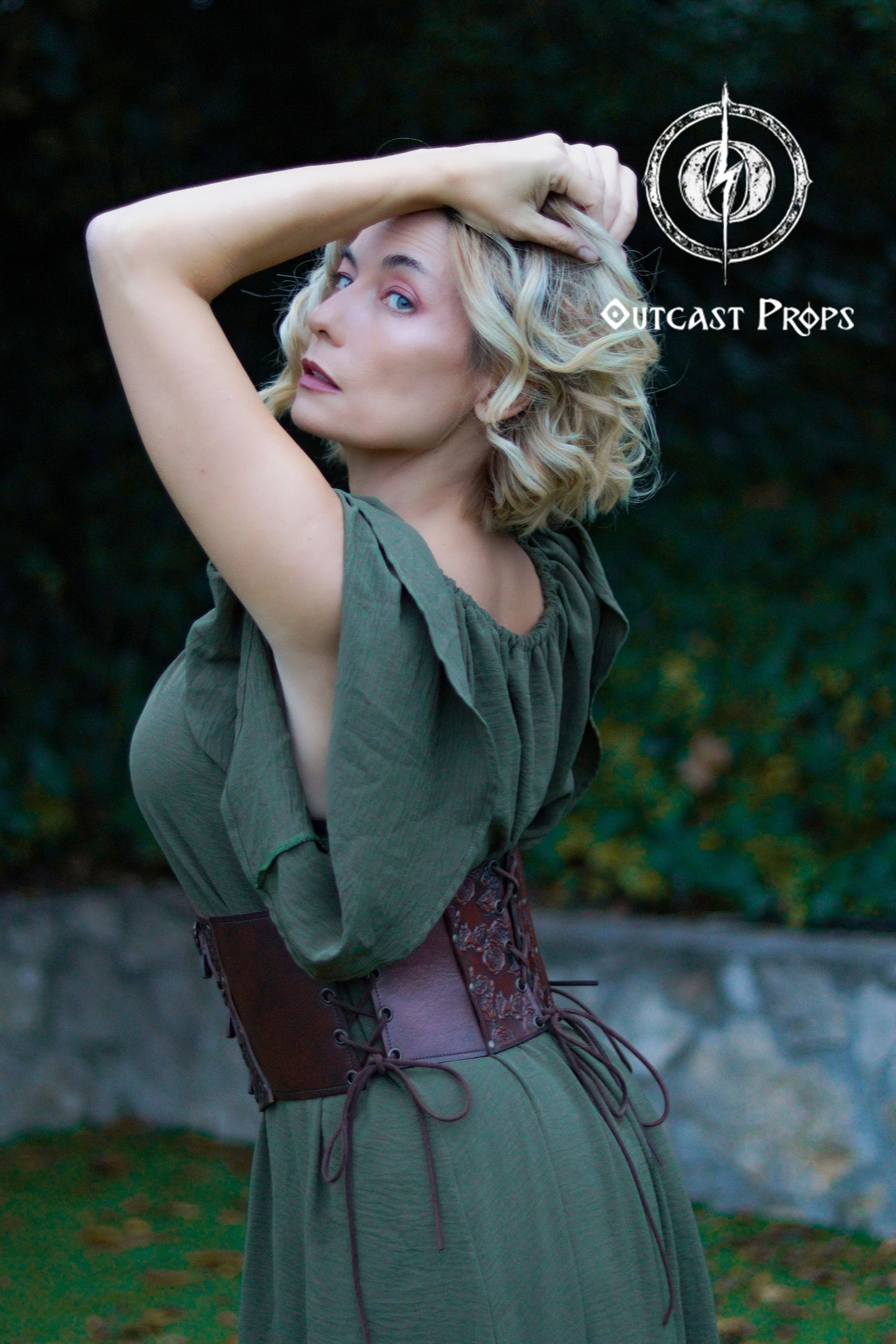 Woman in a green dress with a corset belt posing outdoors, with 'Outcast Props' logo in the corner. Elegant Floral Underbust Leather Corset - Handcrafted Rose Design for Larp, Reinassande Faire &amp; Cosplay
