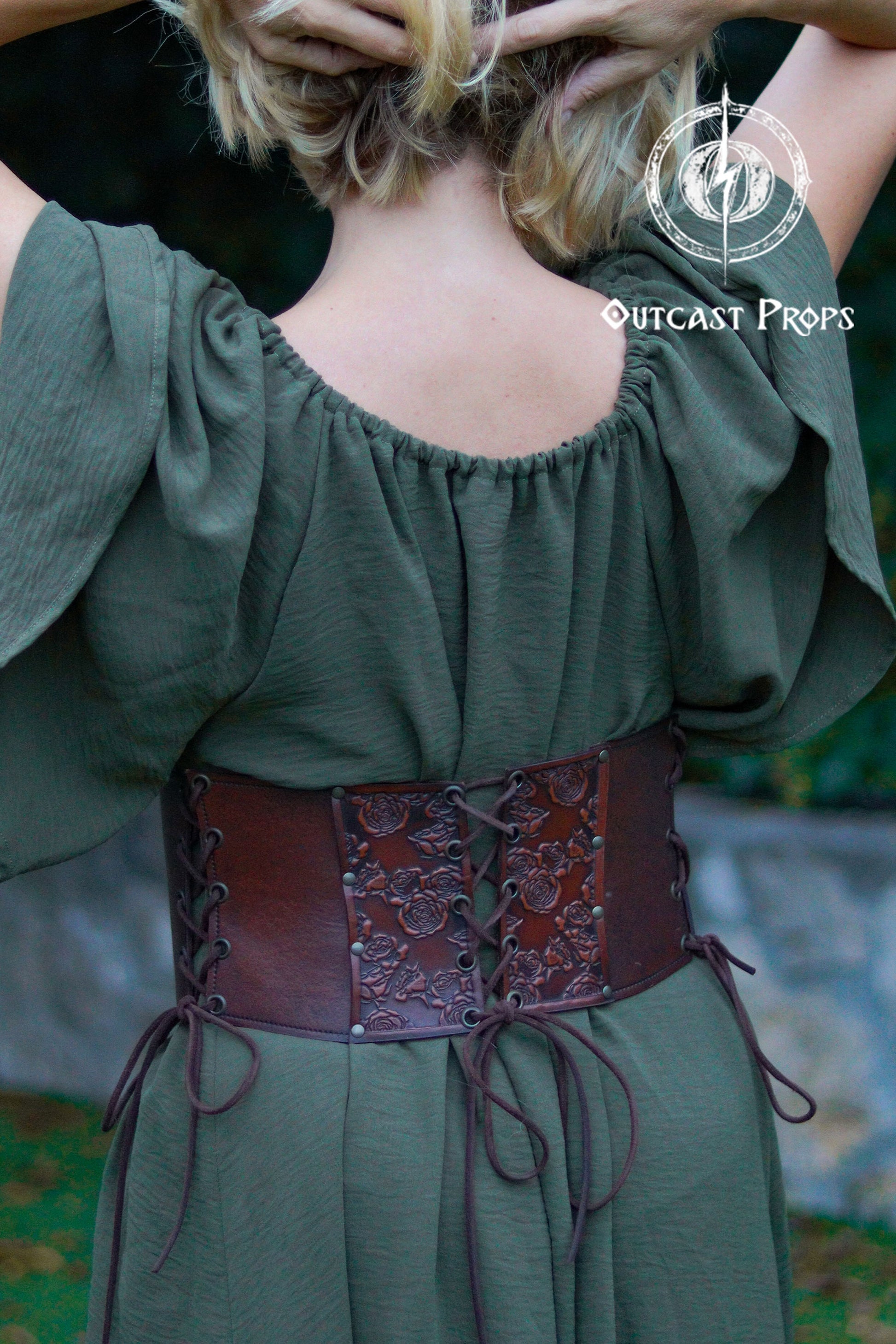 Person wearing a green off-shoulder top with a brown leather belt, brand 'Outcast Props' visible. Elegant Floral Underbust Leather Corset - Handcrafted Rose Design for Larp, Reinassande Faire &amp; Cosplay