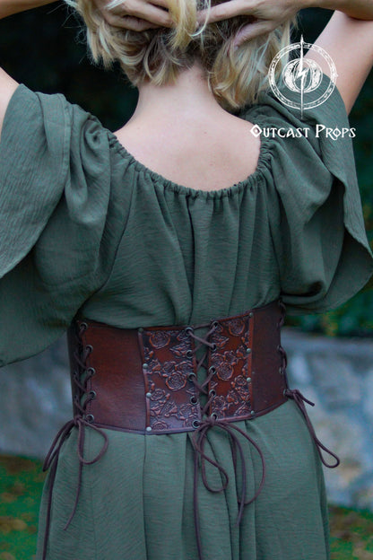 Person wearing a green off-shoulder top with a brown leather belt, brand 'Outcast Props' visible. Elegant Floral Underbust Leather Corset - Handcrafted Rose Design for Larp, Reinassande Faire &amp; Cosplay