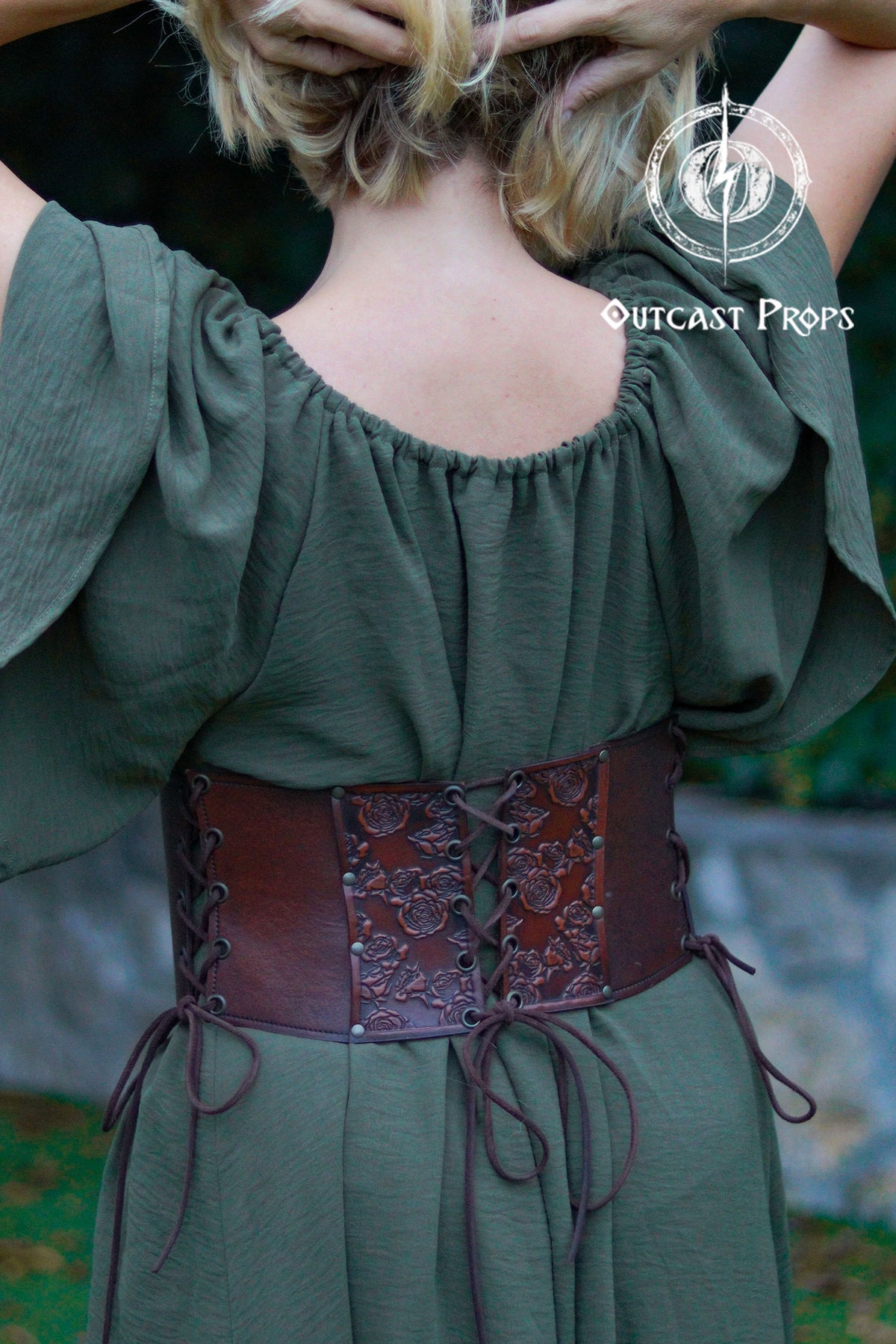 Person wearing a green off-shoulder top with a brown leather belt, brand 'Outcast Props' visible. Elegant Floral Underbust Leather Corset - Handcrafted Rose Design for Larp, Reinassande Faire &amp; Cosplay