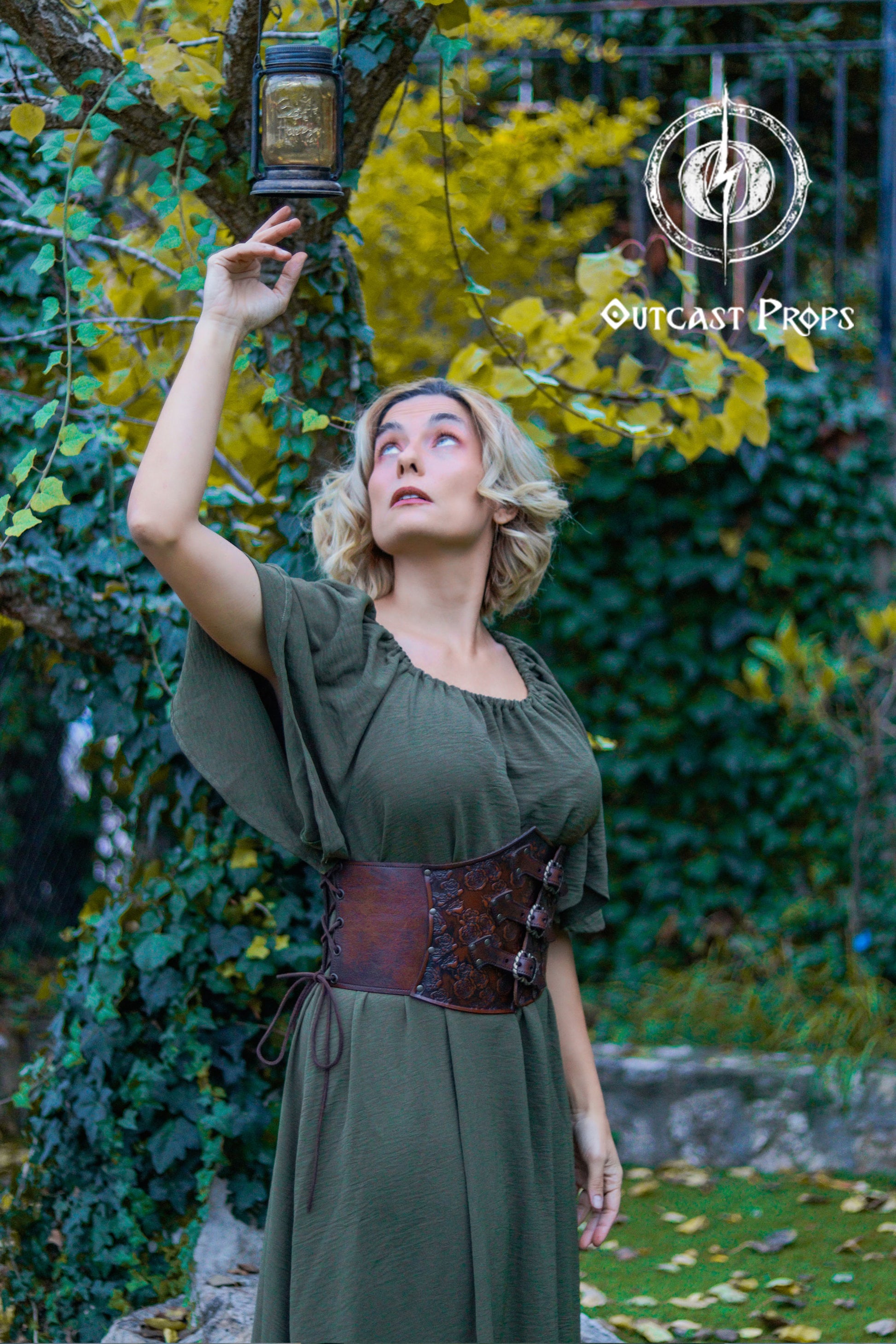 Person in a green dress with a brown belt holding a lantern in a forest setting, with 'Outcast Props' branding. Elegant Floral Underbust Leather Corset - Handcrafted Rose Design for Larp, Reinassance Faire Cosplay