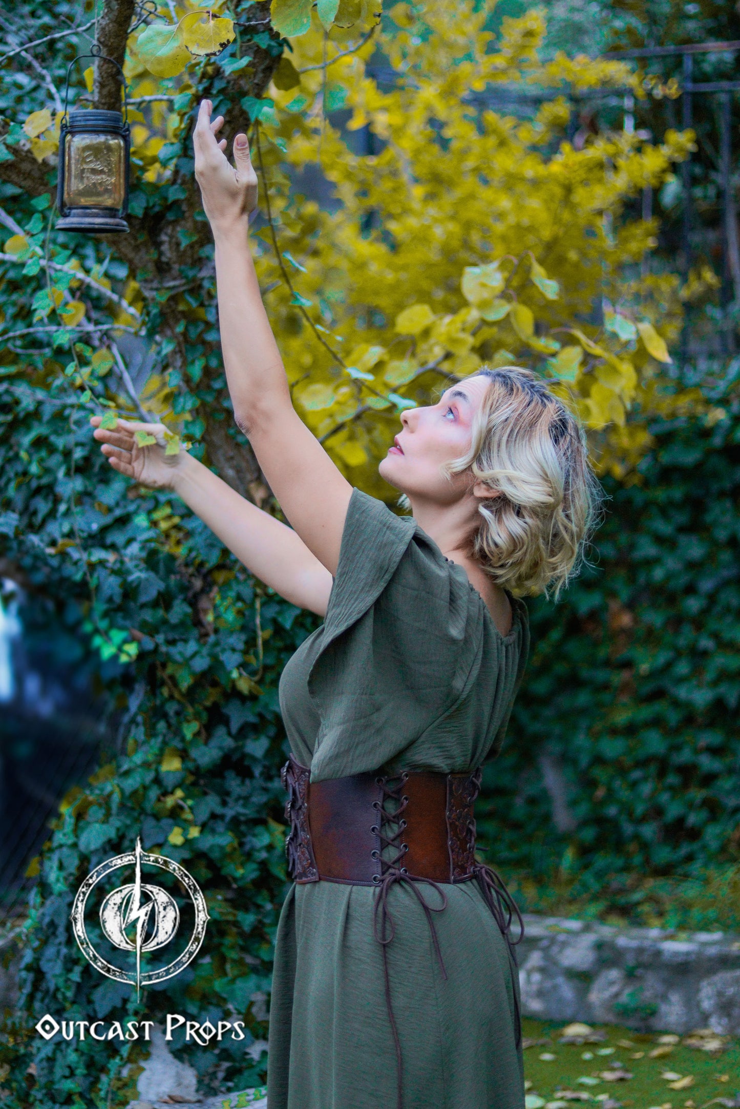 Woman in a green dress reaching up to a tree with a lantern, surrounded by greenery. Elegant Floral Underbust Leather Corset - Handcrafted Rose Design for Larp, Reinassance Faire Cosplay