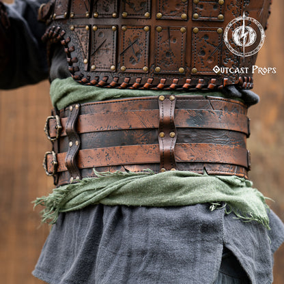 Close-up of a person wearing detailed leather armor with 'Outcast Props' branding. Close-up of a detailed costume with leather straps and a green cloth, branded 'Outcast Props'.  Veteran Leather Belt premium vegetable-tanned, worn by raiders and mercenaries.