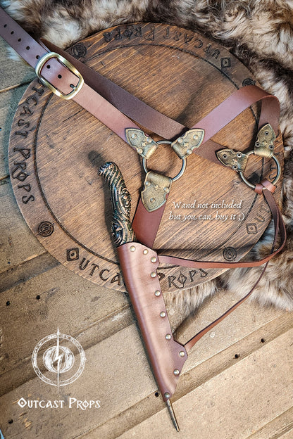 A brown leather wand holster with dual metal rings and riveted construction. Styled for Voldemort-inspired cosplay or LARP magic duels. Crafted for comfort and realism in wizard costumes, especially for Harry Potter fans recreating Death Eater aesthetics.Magic-style leather belt with decorative elements on a wooden surface, featuring Outcast Props branding.