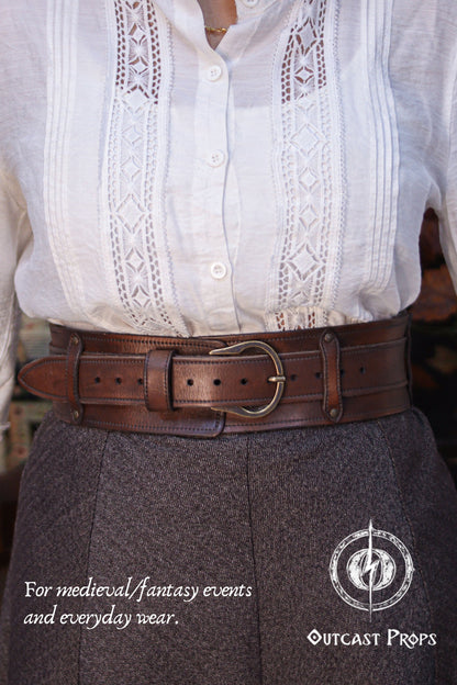 Front view of a model wearing the Handcrafted Leather Wanderer Belt in aged brown vegetable-tanned leather over high-waisted trousers and a white medieval blouse. The belt is shown with the wide outer structure and inner strap aligned, secured by a solid brass buckle. Visible stitching, burnished edges, and natural leather grain highlight artisanal craftsmanship. Modular leathePerson wearing a white blouse with lace details and a brown leather belt, with text about medieval/fantasy events and everyday wear.