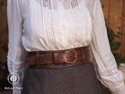 Straight front view of the Handcrafted Leather Wanderer Belt worn high on the waist. The wide leather structure frames the inner belt, showing evenly spaced holes, brass buckle, rivets, and smooth hand-finished edges. Made from premium vegetable-tanned leather with a warm brown aged finish. Designed as a versatile two-in-one belt for LARP, Renaissance fairs, medieval and fantasy cosplay, and practical daily outfits.. Person wearing a white blouse with lace details and a brown belt.