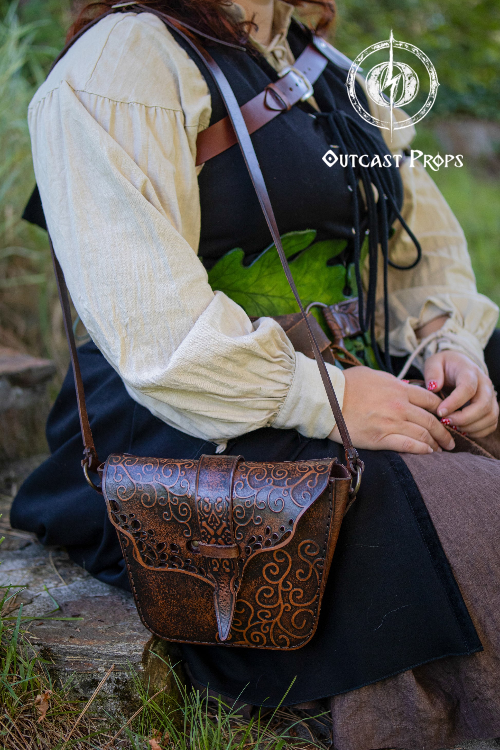 Brown leather witch shoulder bag with embossed floral swirls, worn by a woman in fantasy costume with a green leaf corset and medieval blouse. The bag features aged textures, ornate flap design, and a slim adjustable strap, perfect for renaissance fair outfit, witch costume accessories, LARP shoulder bag, or fantasy leather pouch. The model is seated on a stone step outdoors in a natural setting.