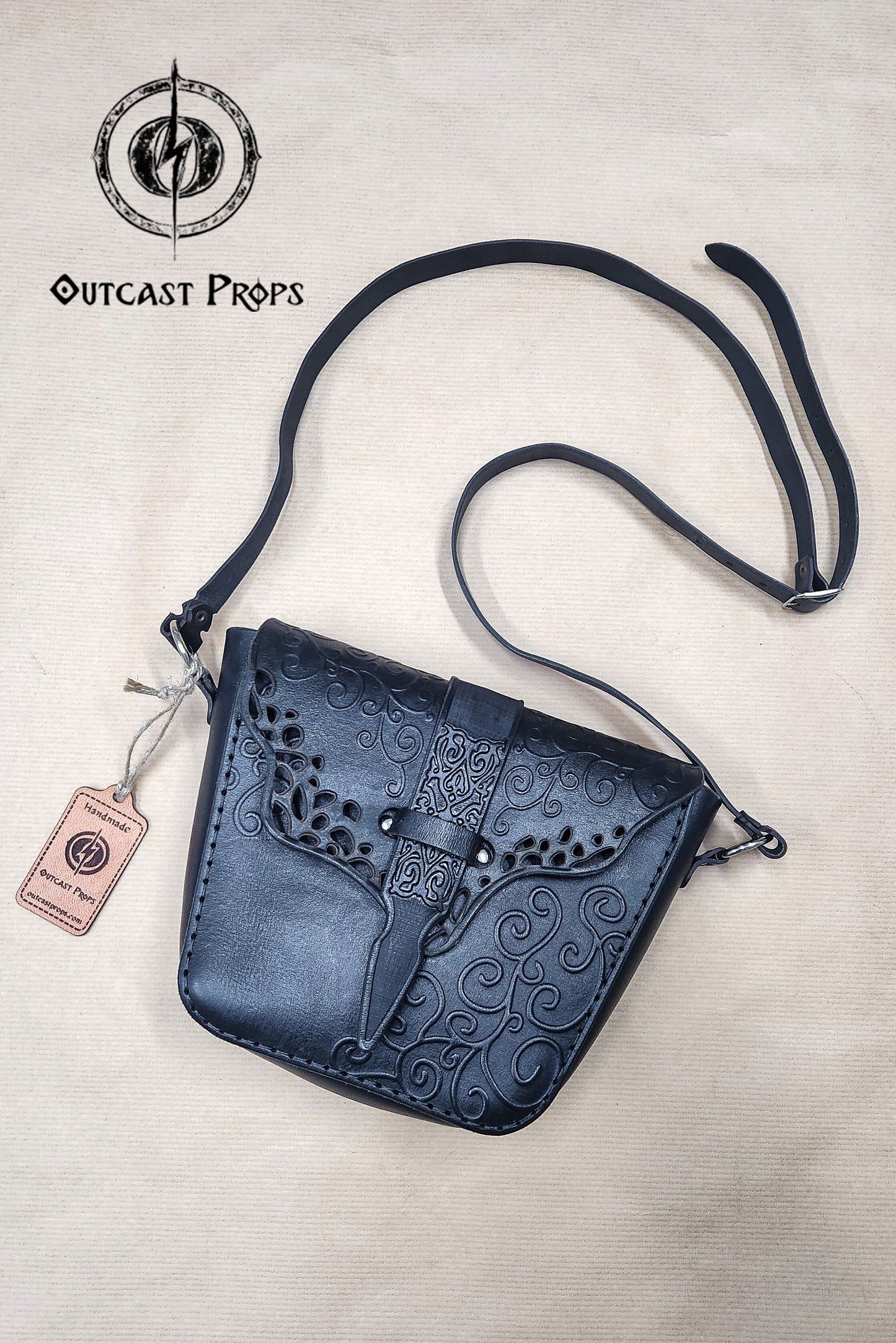 Black leather witch shoulder bag with embossed swirls and intricate leaf pattern, displayed flat on a beige surface with Outcast Props tag and logo. The handmade fantasy leather bag features a long adjustable strap and decorative front flap, ideal for renaissance fair outfit, dark witch accessories, LARP leather pouch, or gothic shoulder bag. High contrast background emphasizes textures and craftsmanship. Gothic Witch Shoulder Bag - Leather Festival Handbag for Dark Fashion Enthusiasts