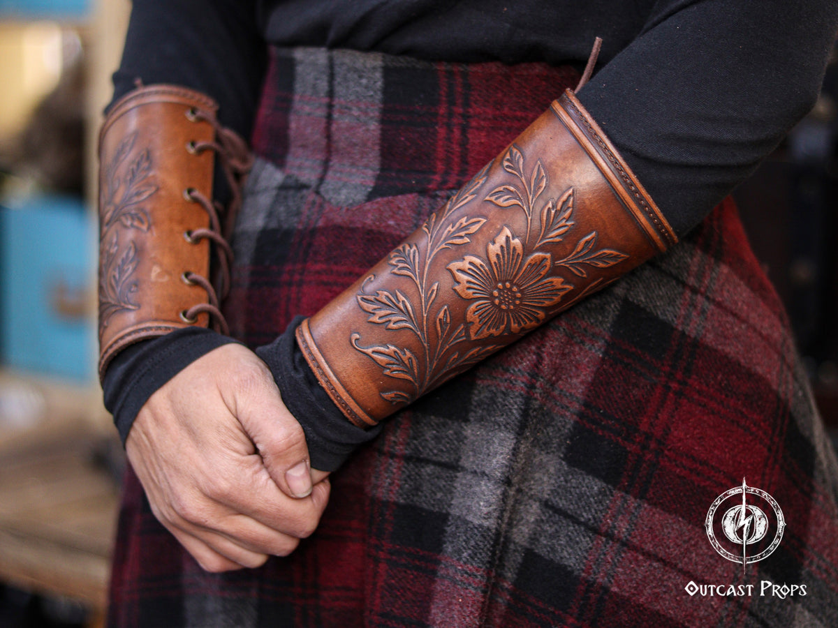 A close up of the Wildflower leather bracer worn on the forearm, showing the detailed floral embossing and aged brown finish. The bracer is laced on the inner side and shaped to fit comfortably on the arm. The person is wearing a dark long sleeve top and a plaid skirt, creating a medieval inspired outfit suitable for renaissance faire, larp cosplay accessory, elven herbalist witch costumes and fantasy themed events.