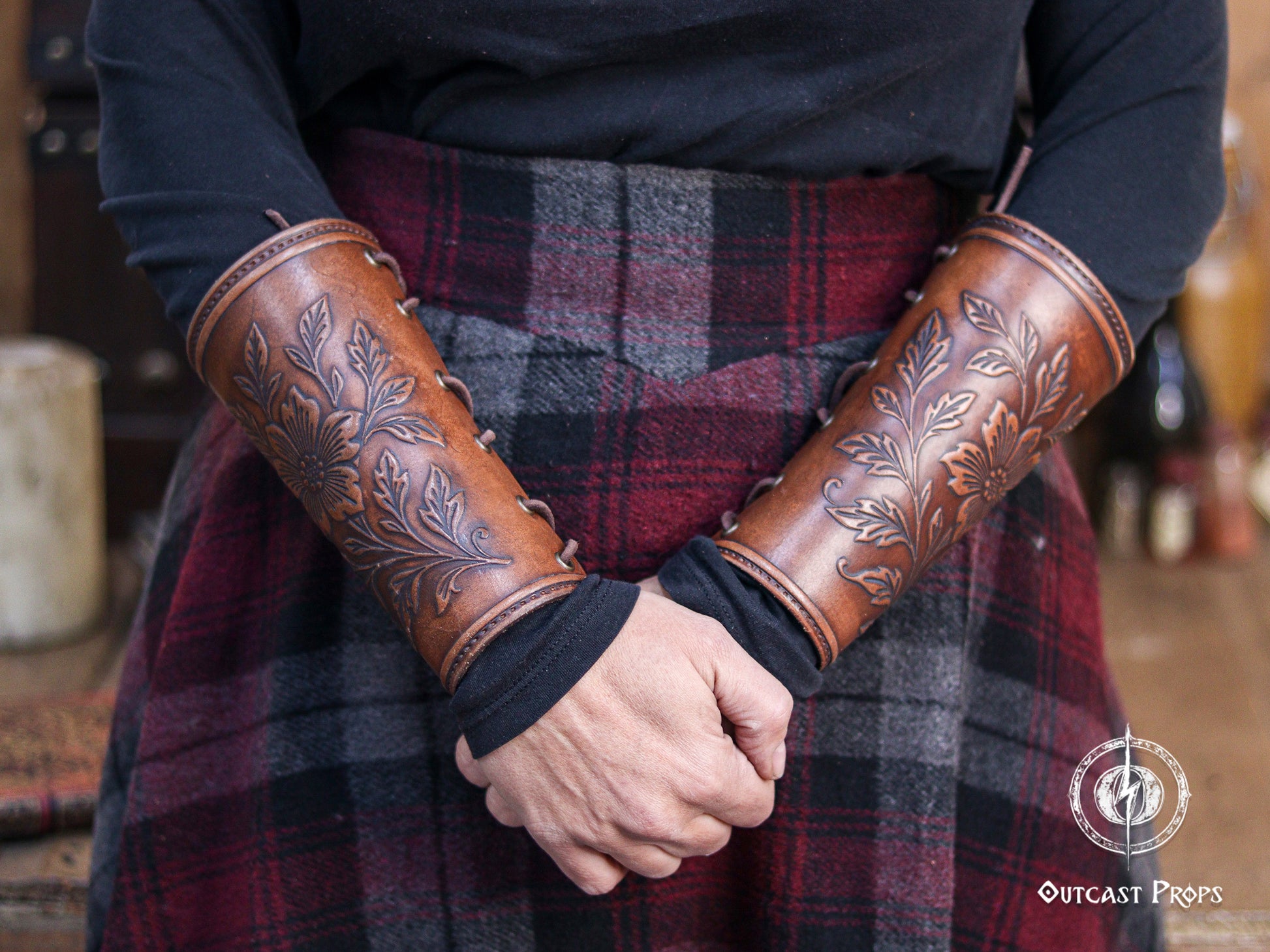 Two Wildflower leather bracers in aged brown are worn on both forearms, showing the floral embossed pattern in full view. The bracers curve naturally around the arm and are paired with a plaid skirt and dark top for a medieval fantasy look. Ideal for renaissance faire outfits, larp cosplay accessory sets, elven characters, herbalists, witches and nature inspired costumes that highlight handcrafted leatherwork.