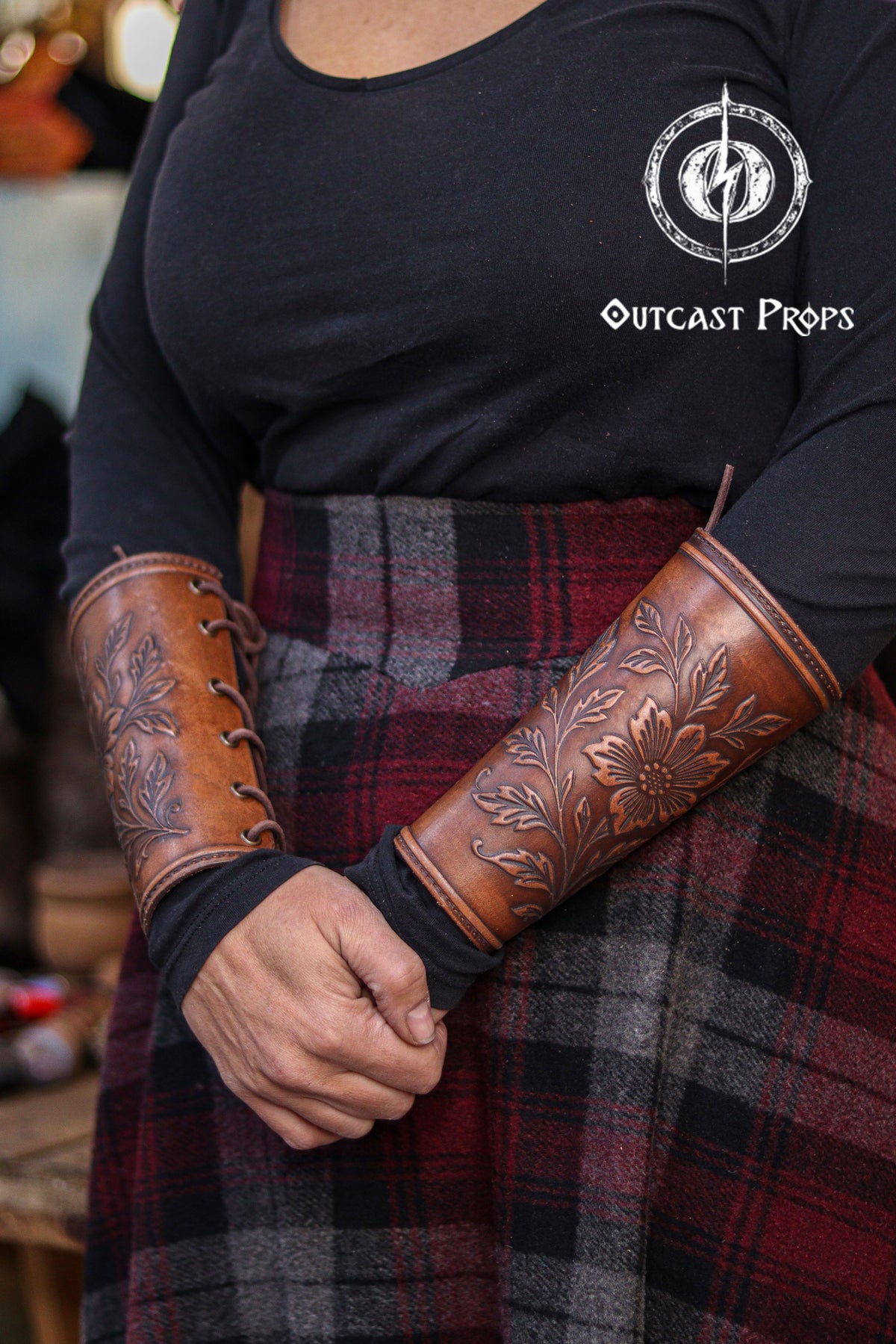 The Wildflower leather bracer is shown worn with a dark long sleeve top and plaid skirt, highlighting its floral embossed design and aged brown tone. The curved shape and lace closure make it a comfortable renaissance faire accessory. Perfect for larp cosplay accessory looks, elven or druid characters, herbalist or witch outfits and fantasy costumes that emphasize natural motifs and handcrafted leather details.