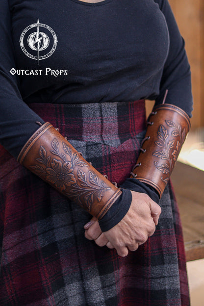 A full view of the Wildflower leather bracers worn on both arms, showing the floral embossed pattern and rich aged brown color. The outfit combines a dark top with a plaid skirt for a rustic medieval style. These bracers work well for renaissance faire costumes, larp cosplay accessory looks, elven characters, herbalists, witches and any fantasy role that benefits from elegant handcrafted leather gear with botanical details.