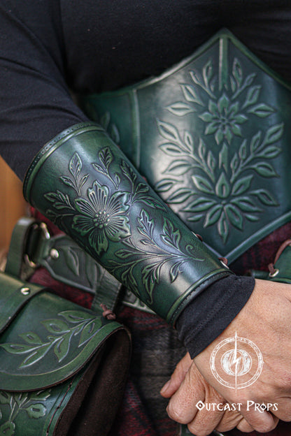 A close view of the Wildflower leather bracer in aged green worn with a matching embossed corset, showing detailed floral patterns on both pieces. The coordinated botanical design creates a natural fantasy aesthetic perfect for renaissance faire looks, larp cosplay accessory sets, elven herbalist witch costumes and nature inspired characters. The image highlights the smooth curve of the bracer and the richness of the green dye.