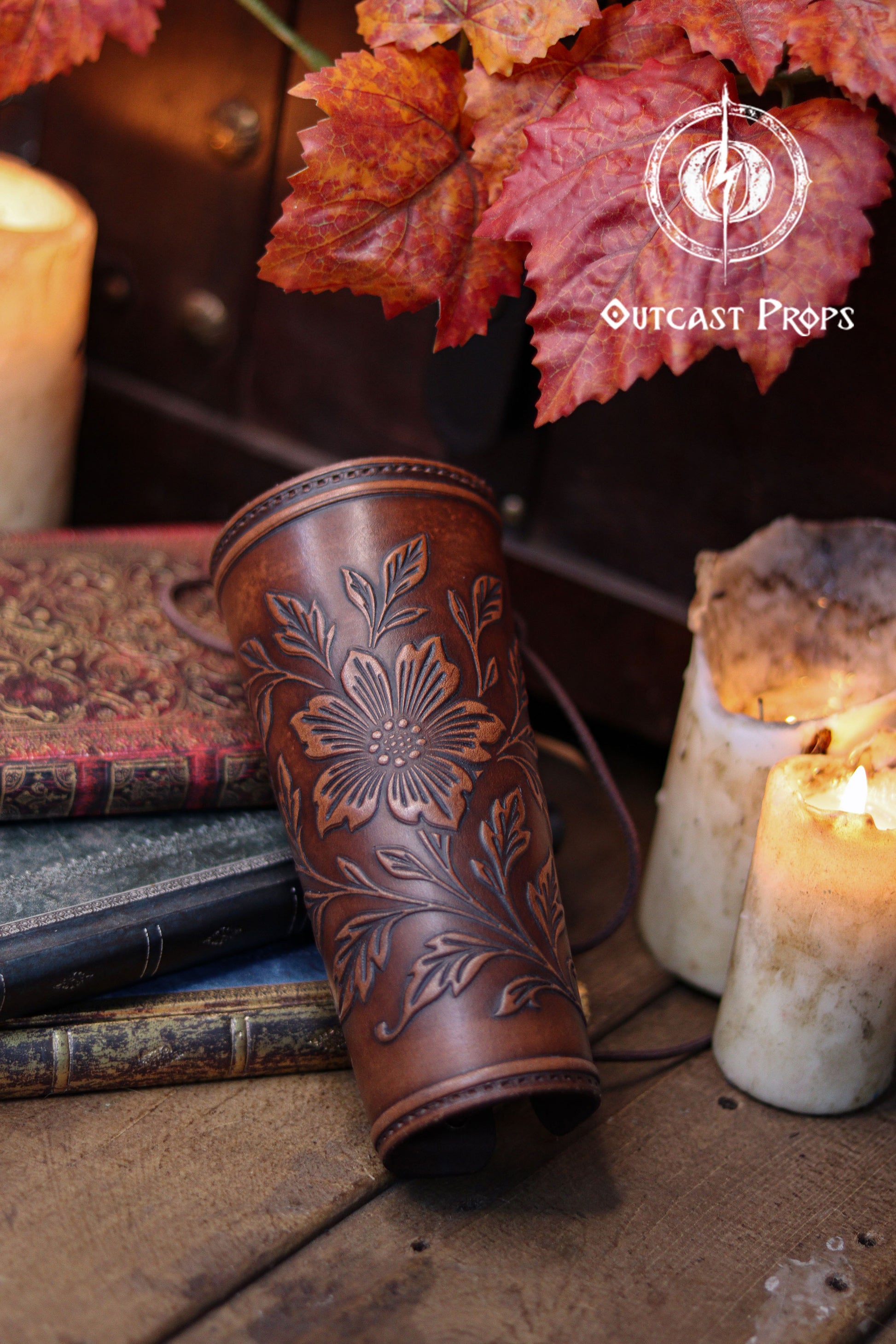 The Wildflower leather bracer in aged brown rests on old books beside warm candles and autumn leaves, showing its floral embossed design in clear detail. The natural texture of the handcrafted leather stands out in this renaissance faire accessory. Perfect for larp cosplay accessory sets, elven herbalist witch characters and nature themed fantasy outfits. The scene highlights the refined botanical pattern and artisanal quality of the bracer.