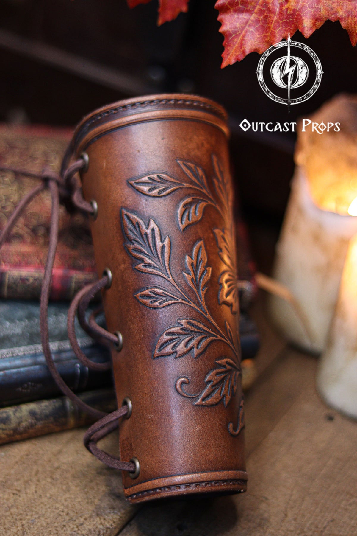 The back view of the Wildflower leather bracer shows the laced closure and the aged brown finish, resting against old books, candles and autumn leaves. This renaissance faire accessory highlights the handmade construction and comfortable fit. Suitable for larp cosplay accessory outfits, elven herbalist witch characters and anyone seeking botanical themed leather gear with elegant embossed details in a fantasy inspired setting.
