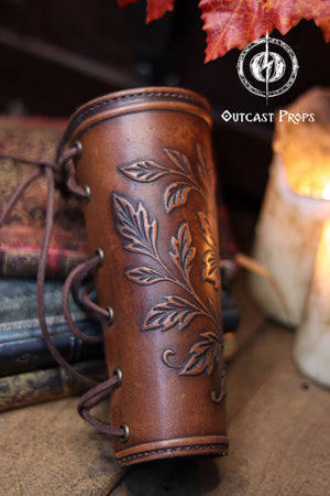 The back view of the Wildflower leather bracer shows the laced closure and the aged brown finish, resting against old books, candles and autumn leaves. This renaissance faire accessory highlights the handmade construction and comfortable fit. Suitable for larp cosplay accessory outfits, elven herbalist witch characters and anyone seeking botanical themed leather gear with elegant embossed details in a fantasy inspired setting.