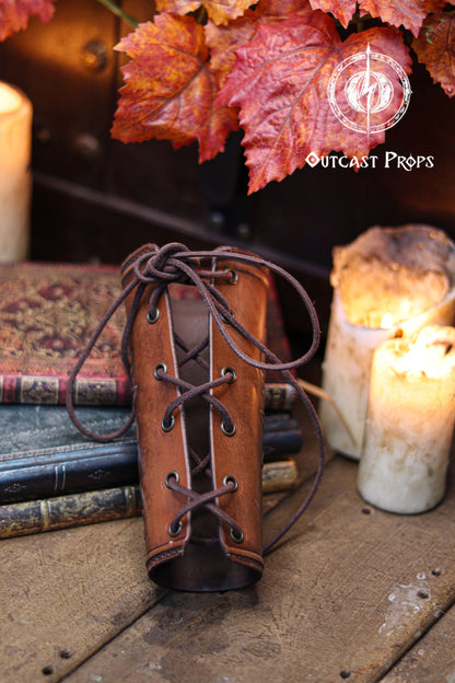 The back view of the Wildflower leather bracer shows the laced closure and the aged brown finish, resting against old books, candles and autumn leaves. This renaissance faire accessory highlights the handmade construction and comfortable fit. Suitable for larp cosplay accessory outfits, elven herbalist witch characters and anyone seeking botanical themed leather gear with elegant embossed details in a fantasy inspired setting.