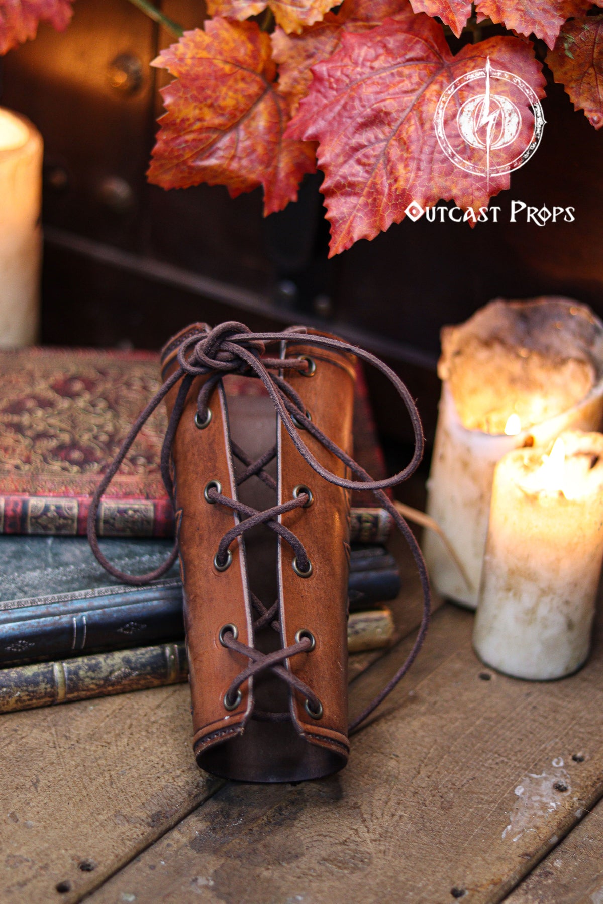 The back view of the Wildflower leather bracer shows the laced closure and the aged brown finish, resting against old books, candles and autumn leaves. This renaissance faire accessory highlights the handmade construction and comfortable fit. Suitable for larp cosplay accessory outfits, elven herbalist witch characters and anyone seeking botanical themed leather gear with elegant embossed details in a fantasy inspired setting.