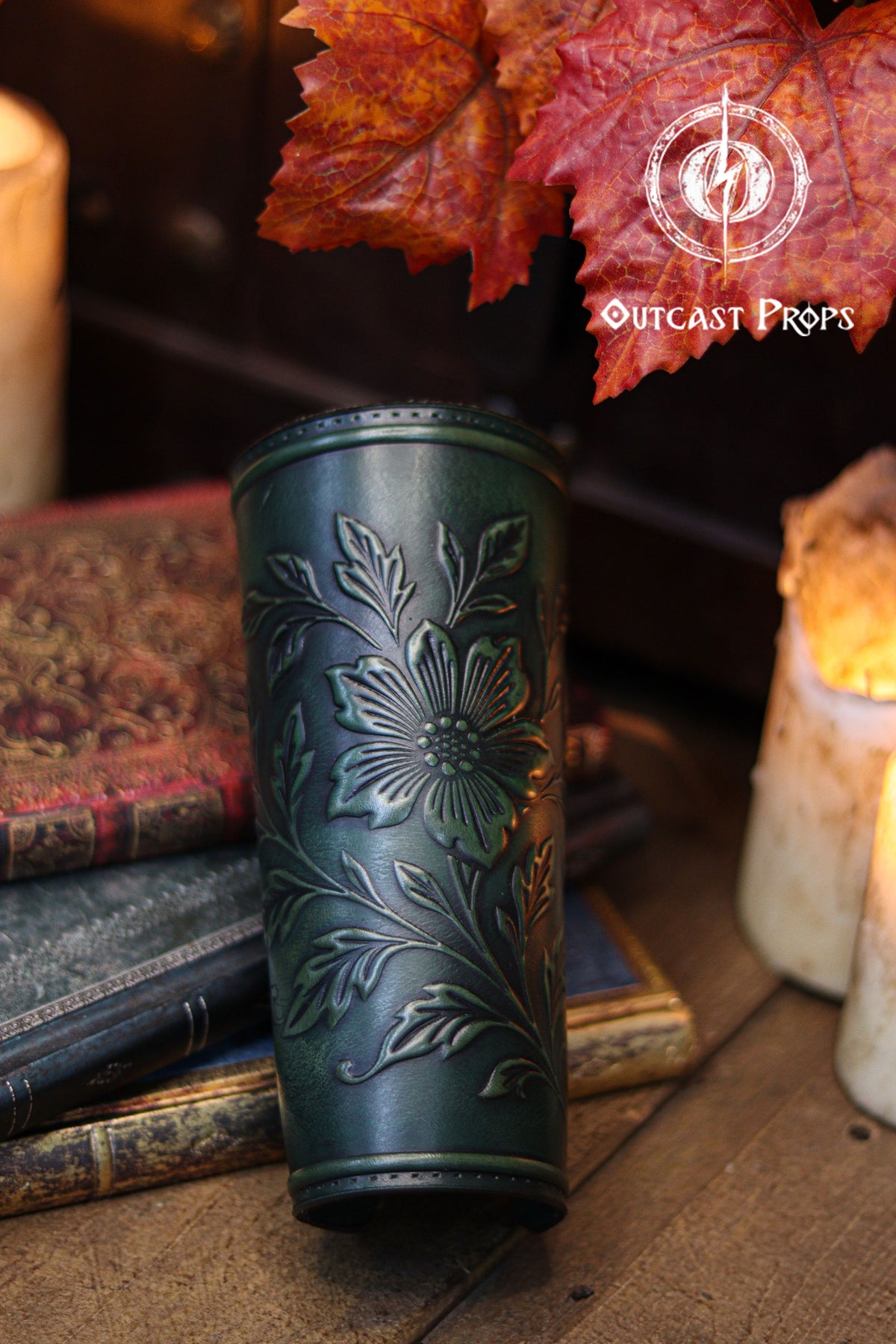 The Wildflower leather bracer in aged green is displayed on stacked vintage books beside candles and autumn leaves. The floral embossed pattern appears vivid against the deep green dye, creating a natural aesthetic perfect for renaissance faire looks. Ideal for larp cosplay accessory sets, elven characters, herbalists, witches and fantasy nature themed costumes where handcrafted botanical leatherwork enhances the outfit.