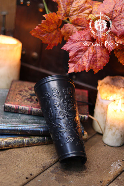 The Wildflower leather bracer in black rests on old books near candles and autumn leaves, showing its embossed floral design with a bold dark finish. This renaissance faire accessory blends elegance with fantasy style, ideal for larp cosplay accessory outfits, elven herbalist witch characters and nature inspired costumes. The scene highlights the craftsmanship and the contrast between the black leather and the botanical pattern.
