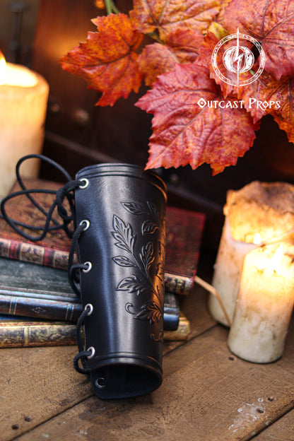 A side view of the Wildflower leather bracer in black shows the laced closure and embossed leaves, placed among vintage books, candles and autumn foliage. This renaissance faire accessory suits larp cosplay accessory ensembles, elven herbalist witch costumes and fantasy themed characters who appreciate detailed botanical leatherwork. The dark finish enhances the floral pattern while maintaining a handcrafted artisan look.