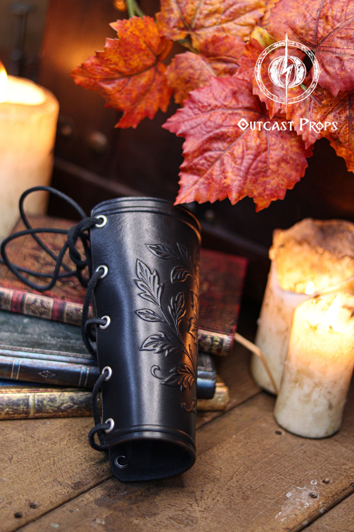 A side view of the Wildflower leather bracer in black shows the laced closure and embossed leaves, placed among vintage books, candles and autumn foliage. This renaissance faire accessory suits larp cosplay accessory ensembles, elven herbalist witch costumes and fantasy themed characters who appreciate detailed botanical leatherwork. The dark finish enhances the floral pattern while maintaining a handcrafted artisan look.